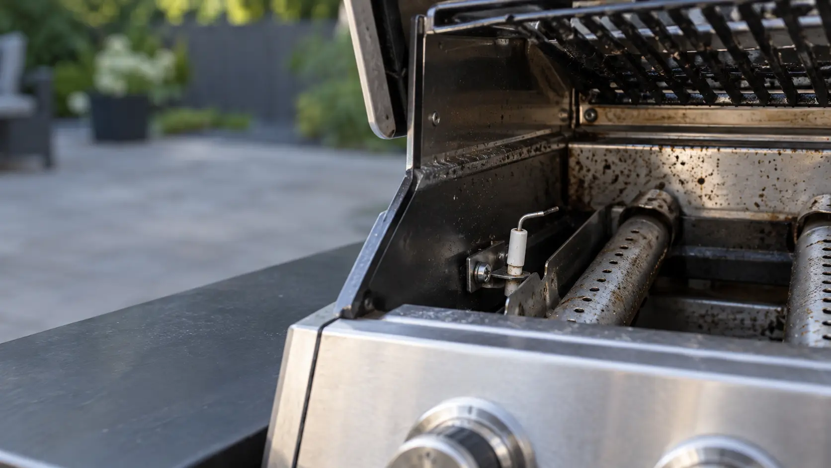 Close view inside the firebox of a cold stainless gas grill with the grate lifted, a white ceramic igniter electrode beside a burner tube, its tip dull with grease and slightly bent away from the ports, fine grease speckles on the metal.