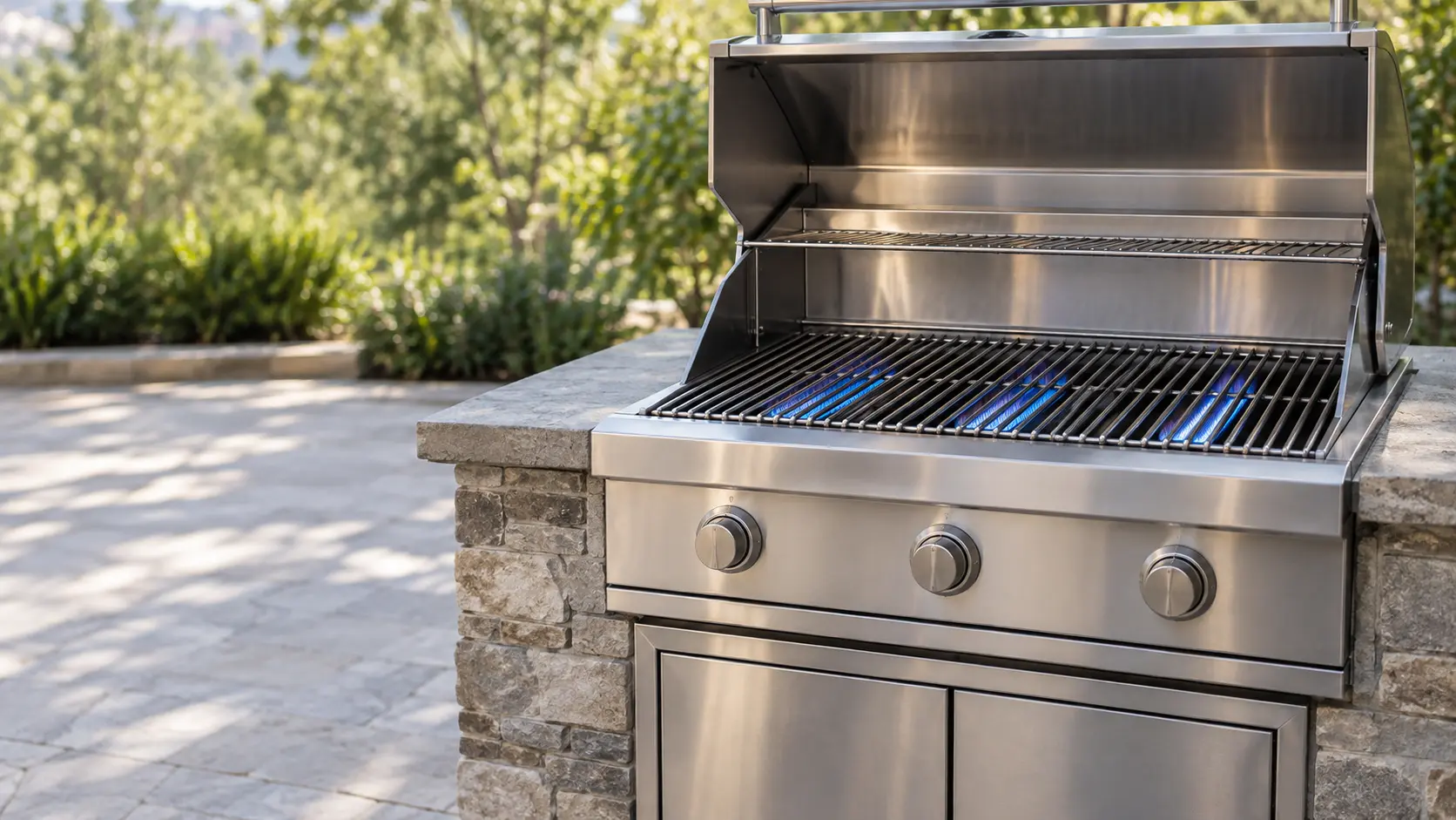 A large stainless built-in gas grill head set into a stone island on a sunny patio, its hood raised to show three burners glowing with even blue flame under clean stainless grates, the knob panel below with knobs, the access doors under the grill closed, plants softly blurred behind, daylight, seasonless.